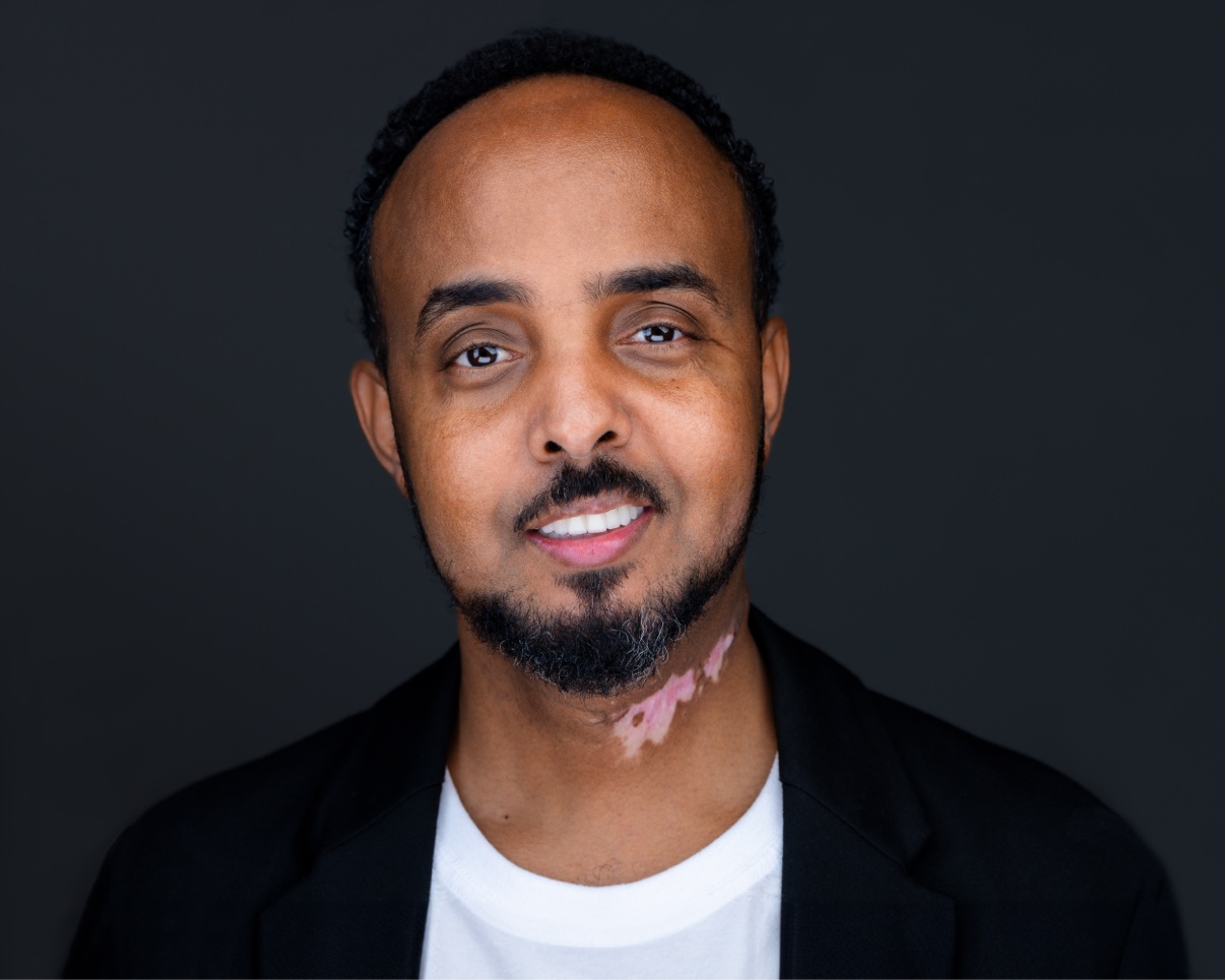 Studio headshot of Musa Aden smiling, in a black blazer and white shirt against a dark background