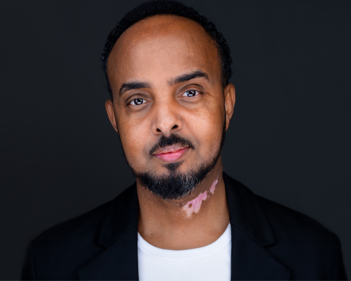 Studio headshot of Musa Aden in a black blazer and white shirt against a dark background
